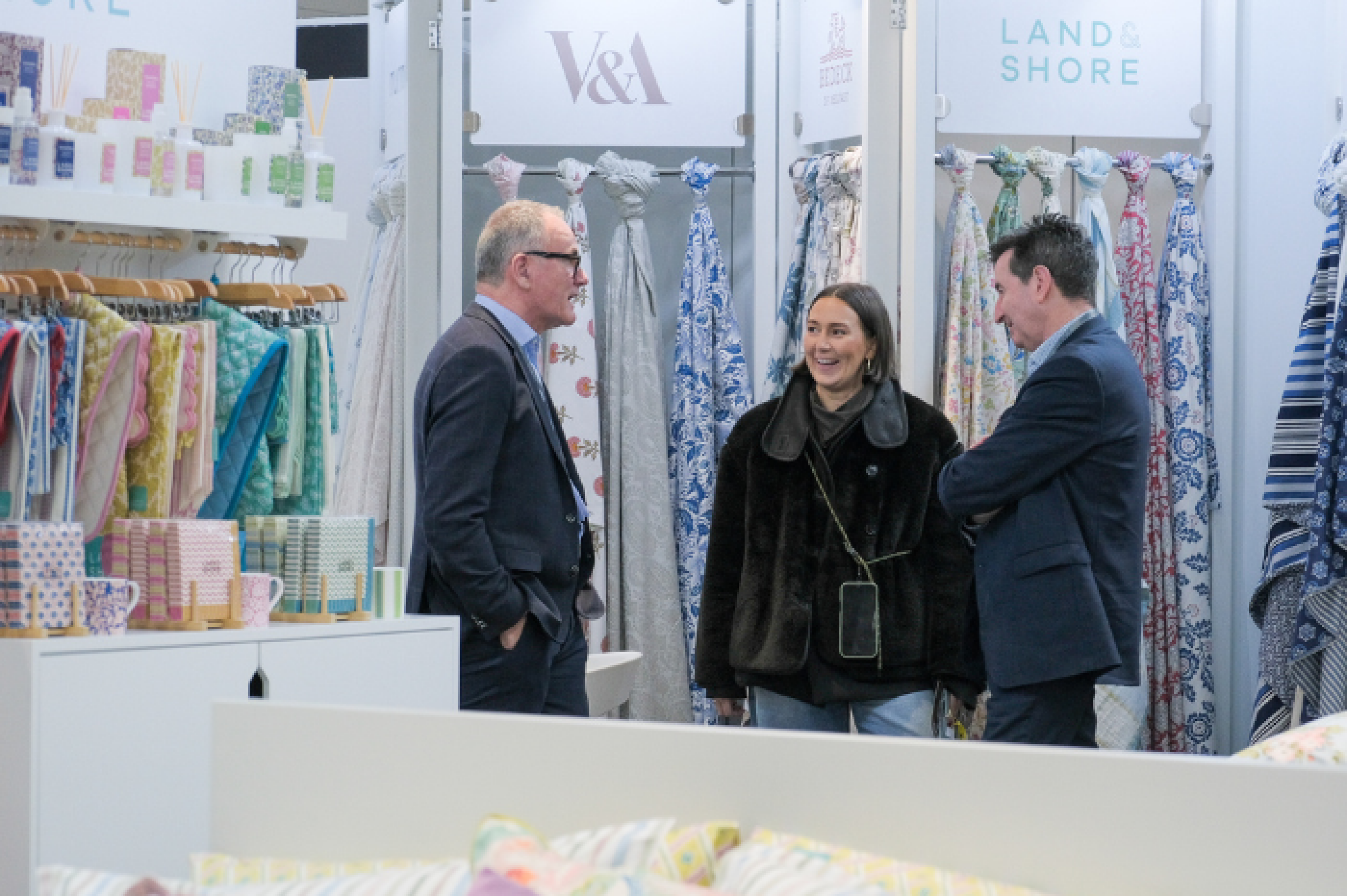 Three people stand central to image at a homewares trade show. Floral and patterned textiles feature behind them in hues of blue and pastel tones all set against a bright white stand with logos V&A, Bedeck, and Land & Shore to top. The Exhibitors and Buyers are chatting and smiling. An image of the top of a bed features to front of image, showcasing a range of linens available to purchase.