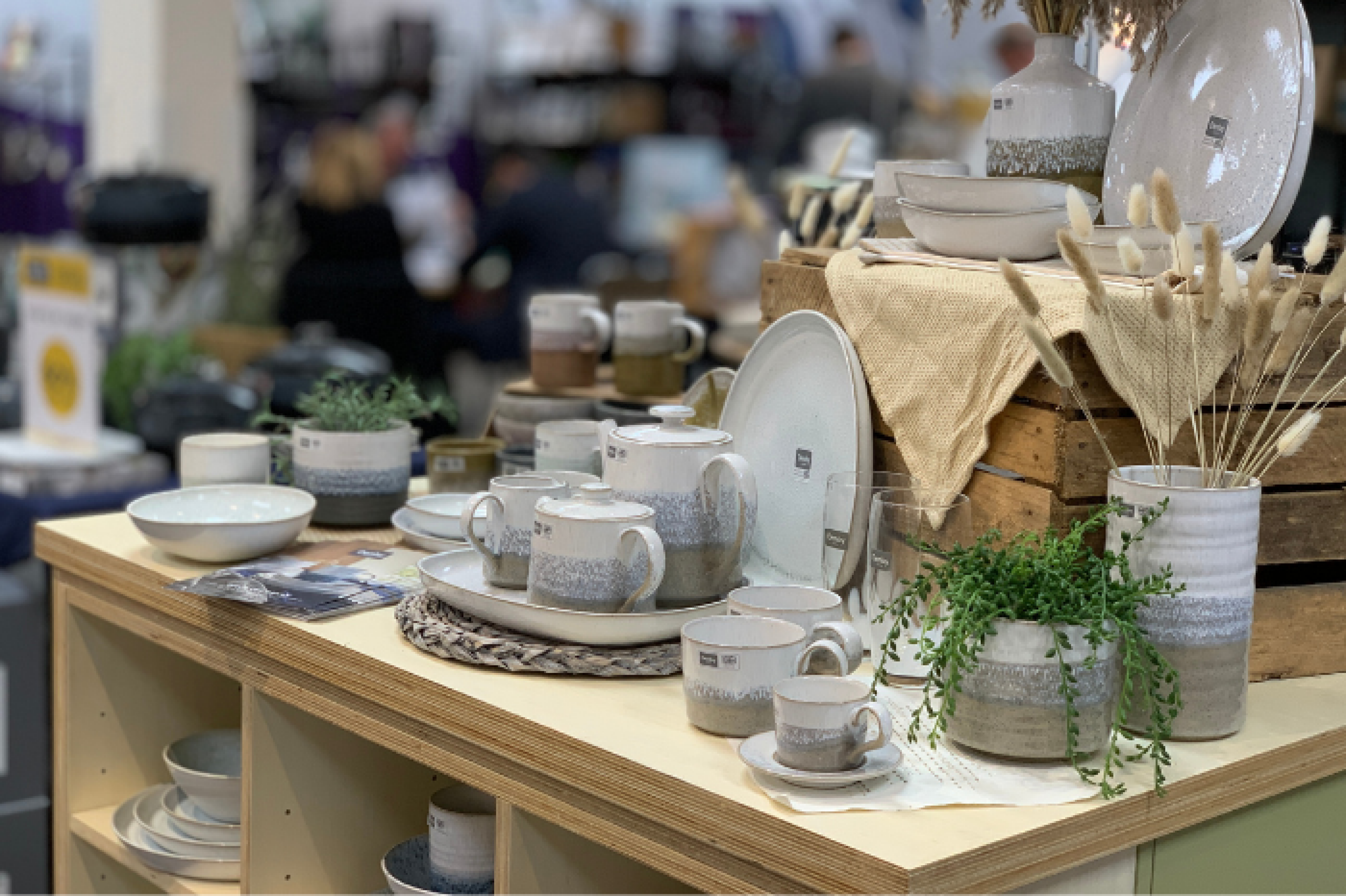 A selection of neutral coloured cermaics are featured on a wooden table, displaying green trailing plants and soft linens at the INDX Home trade show at Cranmore Park