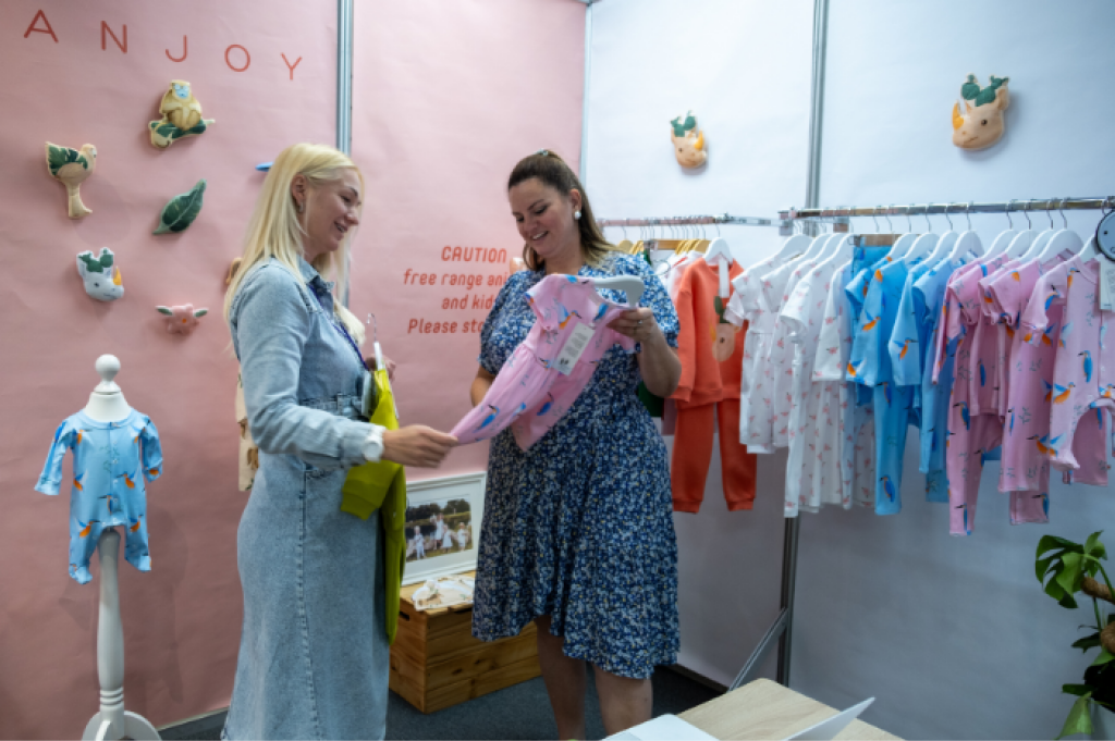 Two women are at the INDX National Kidswear Show, a tradeshow for Indepent Retails at Cranmore Park. In the background there is a peach coloured banner featuring the ANJOY logo and a rail of baby clothing. The two women are smiling and holding a pink short sleeved dress.