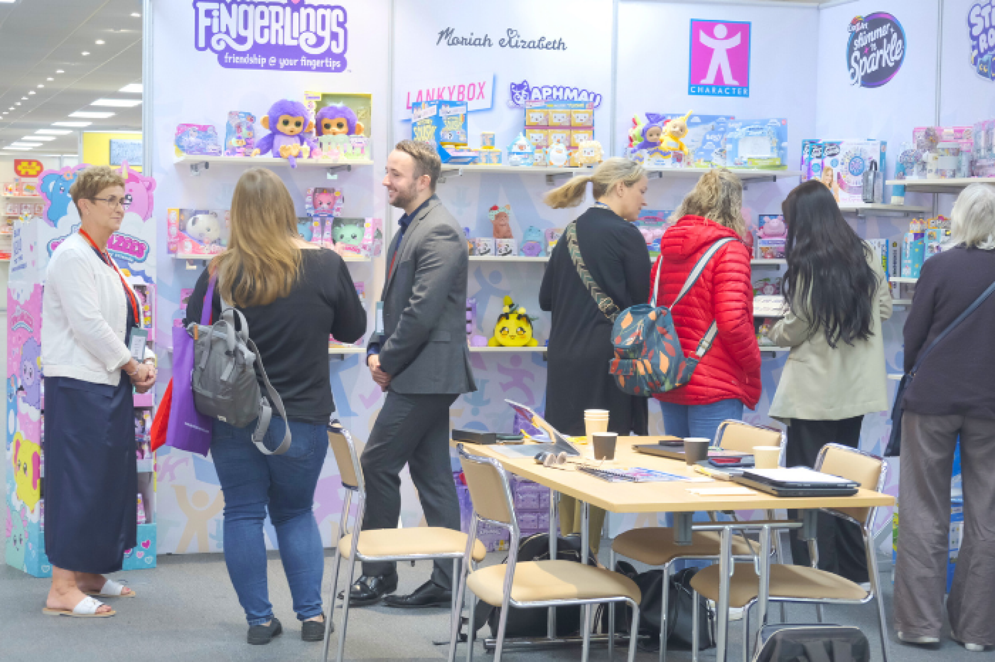 Image of three people networking at INDX Toys - a toy and gift trade show. Bright coloured banners displaying merchandise and logos feature in the background. There are four other Visitors in shot in the background who are shopping the latest toy collections.