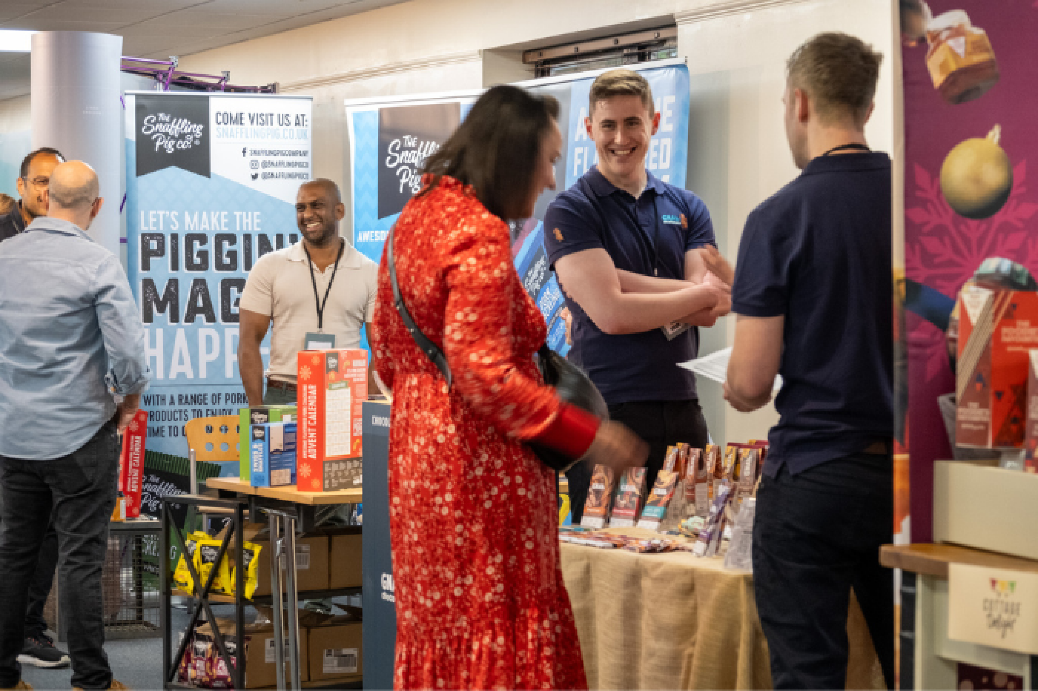 Six people are chatting and doing business at a Gift Food trade show. A woman stands central in a red dress and views a range of chocolate bars from exhibitors Gnaw. The traders are wearing purple polo shiurts and smiling. The Snaffling Pig Co. can be seen trading to the rear of image. The exhibitor is seen smiling at Visitors with a range of crisps, snacks, and scratchings to the front of the stand.
