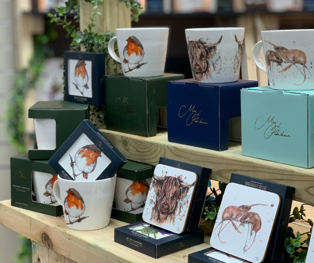 Image from INDX Home trade show at Cranmore Park. The photo features Meg Hawkins mugs and coasters (central) displayed on a wooden table with various nature and animal images on them. There is foliage in the background to add to the homely vibe.