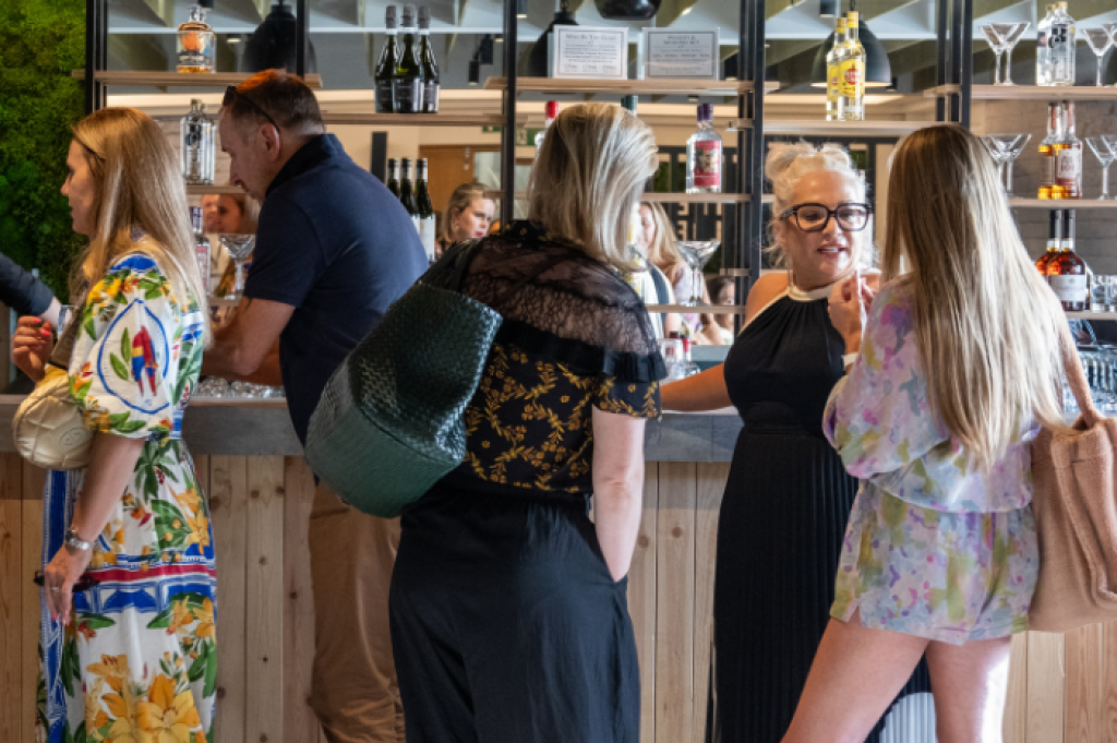 Visitors and key speakers gather at the Summer Soiree event taking place at The Loft at Cranmore Park following the INDX National Intimate Apparel Show. Three women are dressed in evening wear and are standing at the bar chatting and being served cocktails.