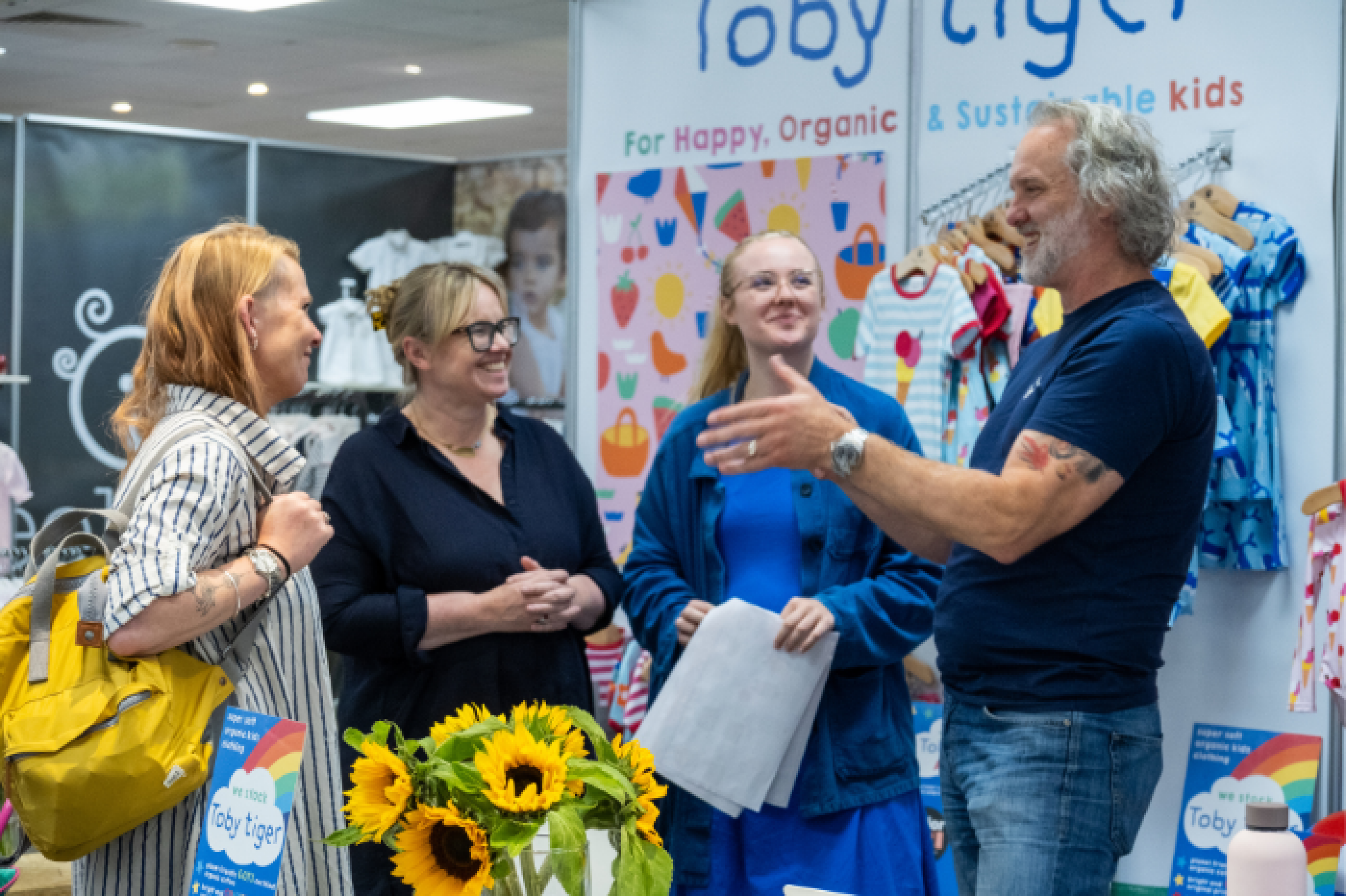 Four people are networking and smiling at INDX National Kidswear Show - a popular tradeshow at Cranmore Park, Solihull. There are stands behind them featuring branded imagery and logos and colour pops of children's clothing. A vase of sunflowers feature in the centre of the image and match the yellow rucksack of an Independent Buyer to the left of the photo.